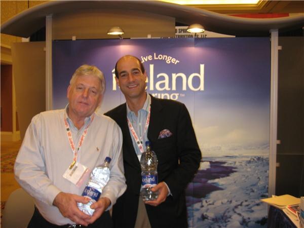 Two men with convention badges each hold a bottle of Iceland water in front of an Iceland Spring backdrop showing an icy landscape.