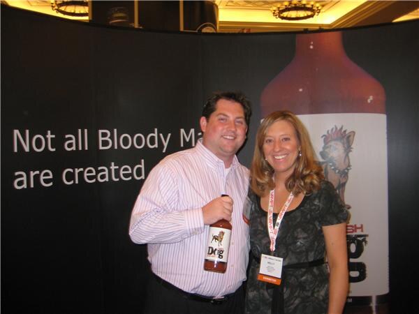 A smiling man holds a bottle of Irish Dog Bloody Mary mix beside a smiling woman wearing a badge, in front of a black backdrop with a large bottle graphic.