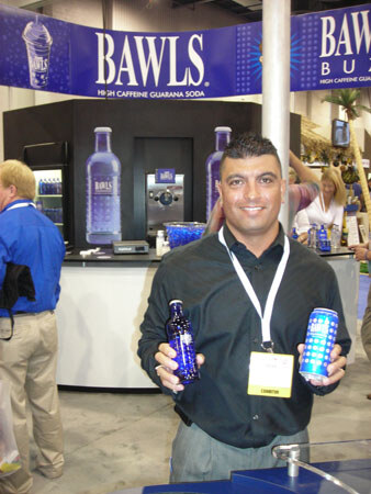 A smiling man in a dark shirt with an exhibitor badge holds a bottle and a can of Bawls in front of a booth with a blue Bawls banner and large bottle graphics.