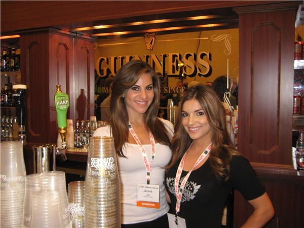 Two smiling women with lanyards stand at a wooden bar with a Guinness sign and a Harp tap handle behind them; Jackie, wearing an exhibitor badge, stands left of center beside stacked cups.