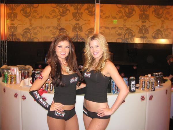 Two women in black Joose outfits stand at a white curved counter displaying Joose cans and cups, in front of a patterned wall.