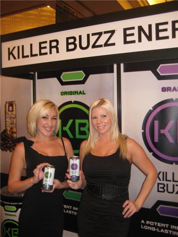 Two blonde women in black dresses hold cans of Killer Buzz Energy in front of a Killer Buzz Energy booth sign with KB logos.