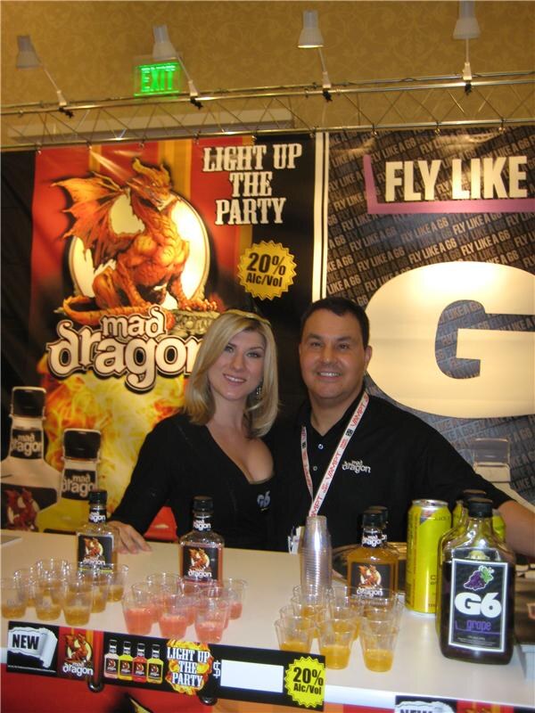 A blonde woman and a man in a black Mad Dragon shirt stand behind a counter with Mad Dragon and G6 bottles and sample cups, in front of Mad Dragon and G6 banners.