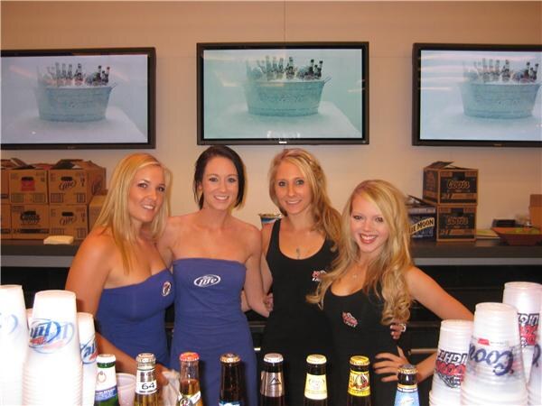 Four smiling women stand behind a counter with beer bottles and plastic cups, two in Miller Lite tops and two in black; screens and Coors boxes are behind them.