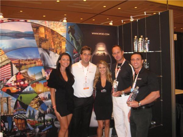 Two women and three men stand together at the NY2O booth; two men hold water bottles, with a New York-themed graphic wall and the NY2O web address behind them.