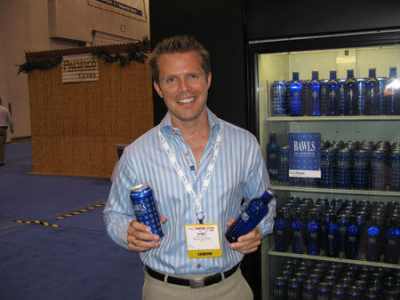 A smiling man in a blue striped shirt holds a blue Bawls can and a blue bottle beside a refrigerated cooler stocked with Bawls Guarana bottles.