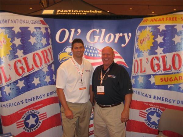 Two smiling men in a booth with Ol' Glory energy drink banners showing Operation Homefront logos. A Nationwide Insurance sign hangs above. One wears a white polo, the other a black shirt.