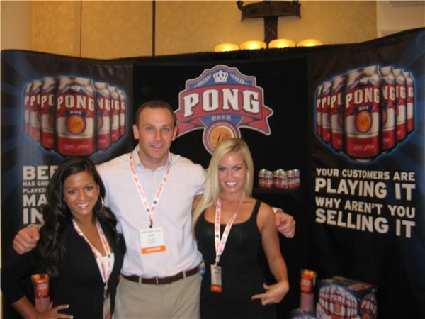A man stands between two women, all smiling and wearing lanyards, in front of a black Pong Beer backdrop showing the logo and red cans of Pong Beer. A sign reads, Your customers are playing it.
