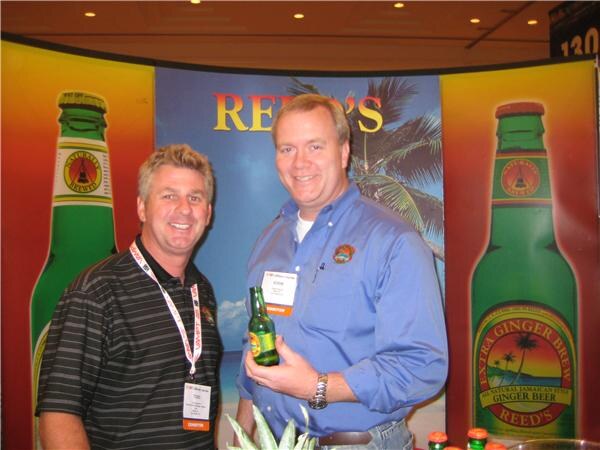 Two smiling men stand in front of a Reed's backdrop with large Extra Ginger Brew bottle graphics. The man on the right, in a blue shirt, holds a green bottle of Reed's ginger beer.