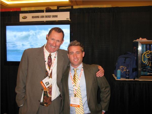Two men in suits and lanyards stand with arms around each other at a Roaring Lion Energy Drink booth with a black curtain, a wave video on a screen, and a small cooler with cans on the right.