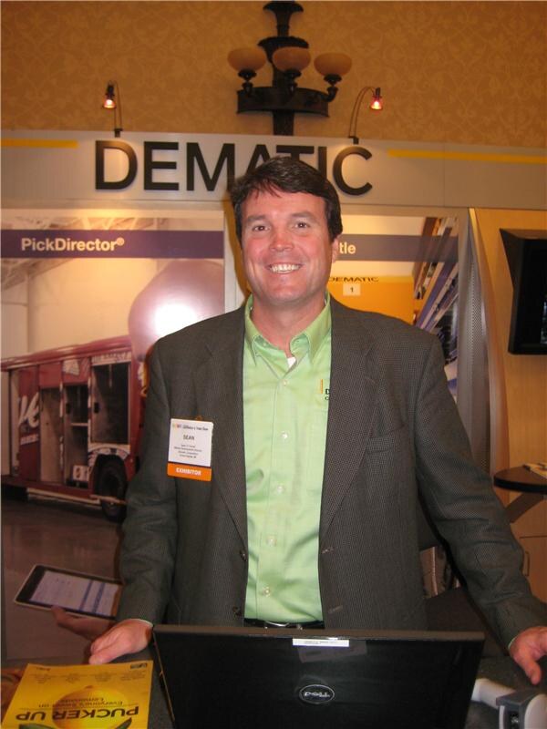 Sean, wearing an exhibitor badge, a green shirt and a tweed jacket, smiles behind a laptop at the Dematic booth, where a PickDirector sign and a delivery truck image are visible.
