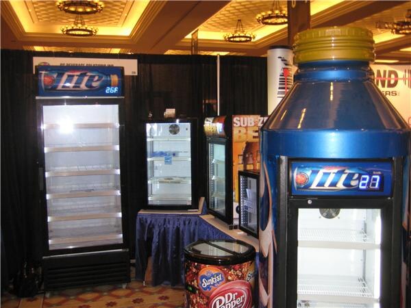 Display of glass-door refrigerators, including Miller Lite coolers, one shaped like a bottle, and a round Sunkist and Dr Pepper cooler in the foreground, in a ballroom with black curtains.