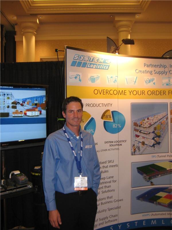 A man in a blue button-up shirt and lanyard stands beside a System Logistics display with charts and warehouse graphics. A screen to the left shows a 3D warehouse model.