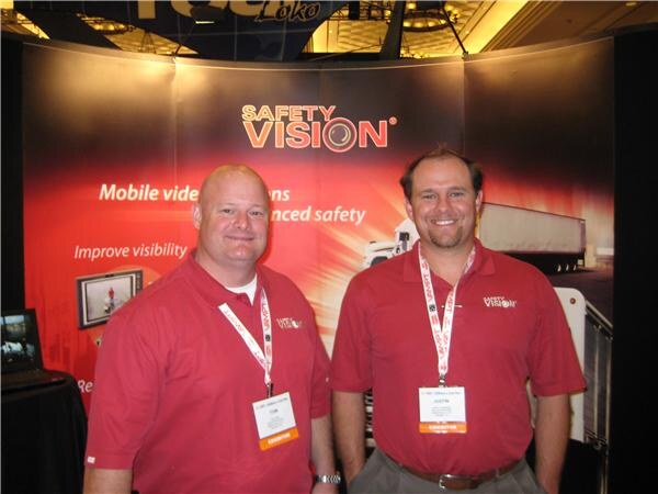 Tom, left, and Justin, right, wear red Safety Vision polo shirts and exhibitor badges in front of a red Safety Vision display about mobile video and enhanced safety.