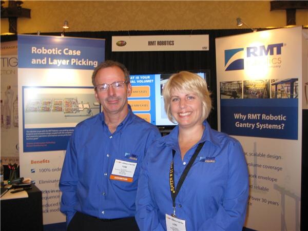 Tom, left, and Lori, right, both in blue RMT Robotics shirts, smile in front of banners about robotic case and layer picking and RMT robotic gantry systems.