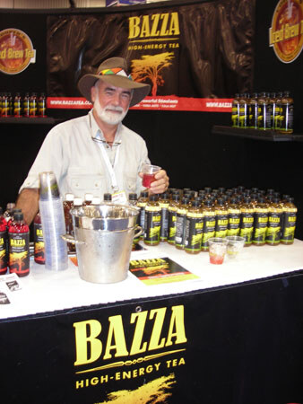 A bearded man in a wide-brimmed hat holds a small cup behind a counter of Bazza High-Energy Tea bottles, an ice bucket and cups, with Iced Brew Tea signs in the background.
