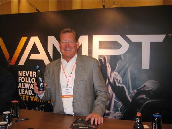 A smiling man in a gray blazer and exhibitor badge holds a bottle of VAMPT and stands behind a table with more VAMPT bottles and cans, in front of a large black VAMPT backdrop.