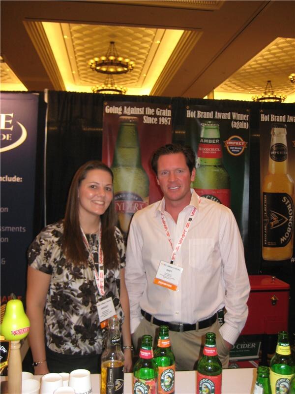 A woman and a man wearing exhibitor badges stand smiling behind a counter of Woodchuck Hard Cider bottles, in front of banners for Woodchuck and Strongbow cider.