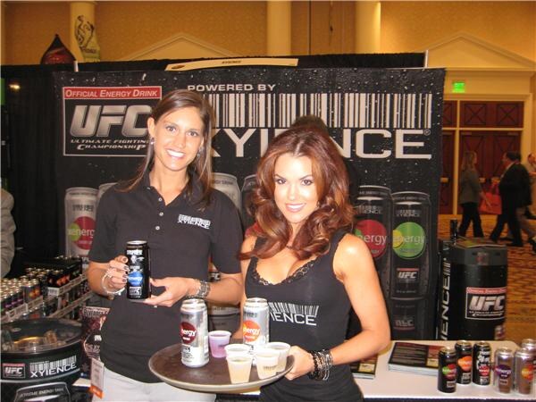 Two smiling women stand at a Xyience booth in front of a UFC and Xyience banner. One holds a can of Xenergy and the other holds a tray with small cups and more cans.