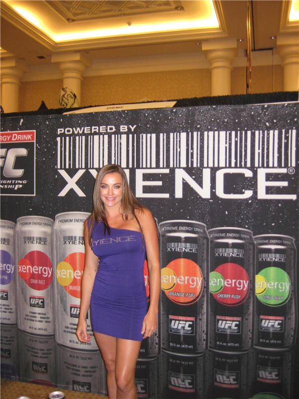 A woman with long brown hair in a purple one-shoulder dress stands in front of a Xyience backdrop showing rows of Xenergy cans in several flavors with UFC logos.
