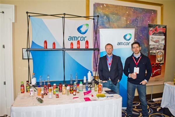 Two men in lanyards stand beside a table of assorted plastic beverage bottles, in front of Amcor banners. A Cold Spring Brewing Company banner stands at the right.