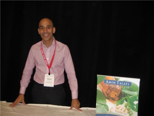 A smiling bald man in a pink shirt and red lanyard stands with his hands on a draped table, next to an Amin Talati Attorneys at Law sign showing herbs and capsules.