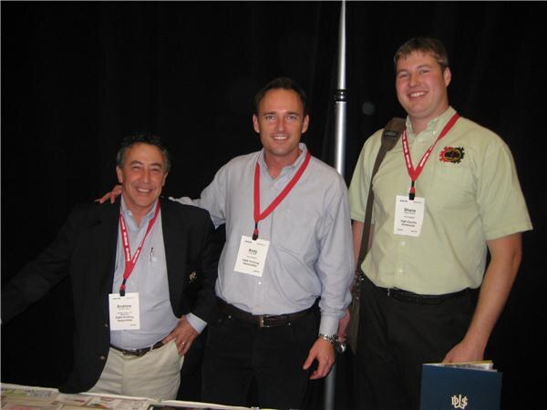 Andrew, Andy and Shane stand side by side behind a table, smiling and wearing red lanyards with name badges, in front of a black curtain.