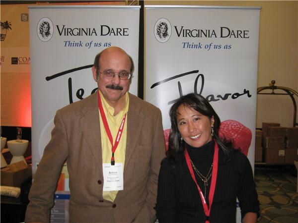 Anton, a man in a brown jacket and yellow shirt with a badge, stands at left beside a smiling woman in black, in front of two Virginia Dare banners.