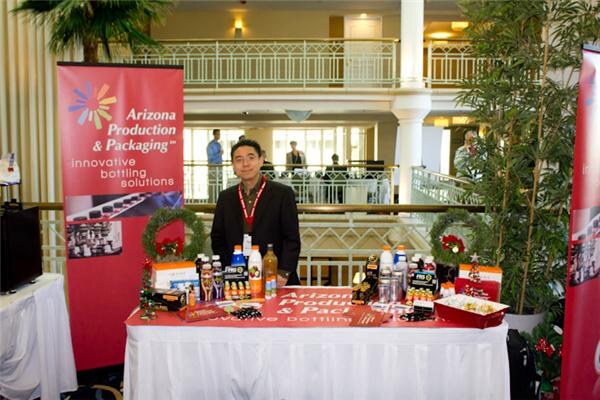 A man in a dark jacket stands behind a table with bottled beverages and holiday decorations, beside red Arizona Production & Packaging banners reading innovative bottling solutions.
