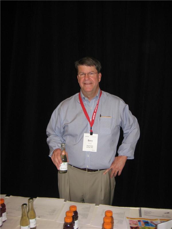 Barry, wearing a badge and red lanyard, stands with one hand on his hip and holds a small glass bottle behind a table with more bottles and papers, in front of a black curtain.