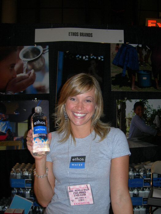 Anne-Michelle Seiler of Ethos Brands smiles and holds a bottle of Ethos Water in front of the Ethos Brands booth sign and photos. Cases of bottled water are stacked behind her.