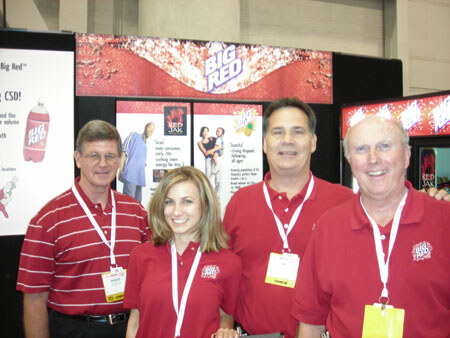 Three men and a woman in red polo shirts and lanyards pose together in front of Big Red and Red Jak banners.