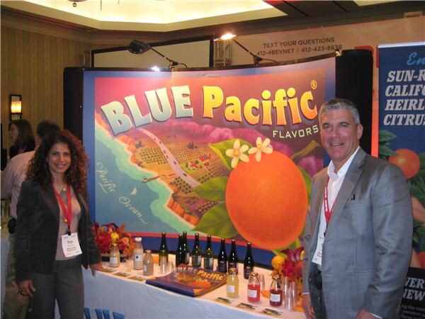 A woman and a man in blazers and lanyards stand on either side of a Blue Pacific Flavors booth. The backdrop shows a large orange, and the table holds bottles and sample jars.