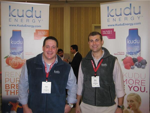 Bob (left) and Nick (right) of Kudu Energy smile in Kudu vests and badges between two Kudu Energy banners showing tangerine and berry blend drinks.
