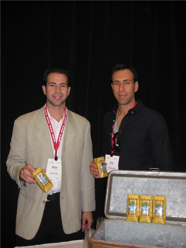 Brian of Coco Cafe, left, and Elan, right, each hold a yellow Coco Cafe carton in front of a black curtain, with more cartons in a metal cooler.