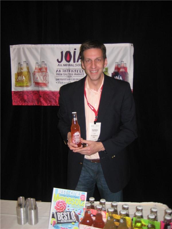 Carleton, wearing a badge and dark blazer, holds a bottle of JOIA all natural soda in front of a JOIA banner, with more bottles and a magazine on the table.