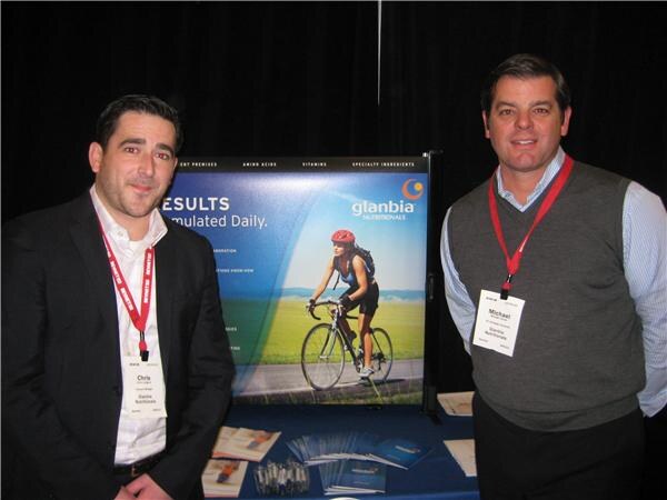 Chris (left) and Michael (right) of Glanbia Nutritionals stand with badges beside a Glanbia Nutritionals banner showing a cyclist, with brochures on the table.