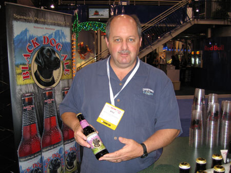 A man in a gray-blue shirt with an exhibitor badge holds a bottle of Black Dog Ale beside a Black Dog Ale banner showing a black dog. Plastic cups are stacked to the right.
