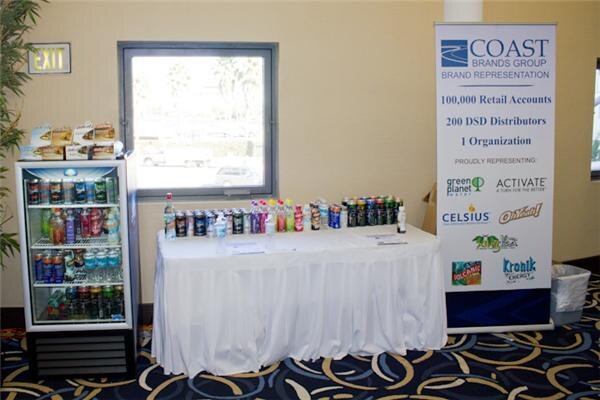 A table with a white skirt holds beverage bottles and cans beside a small cooler and a Coast Brands Group banner listing Green Planet, Activate, Celsius, Oh Yeah! and Kronik Energy.