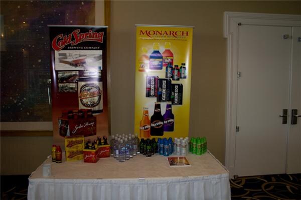 A draped table with Cold Spring Brewing Company and Monarch Custom Beverages banners, with bottled water, canned and bottled drinks and a brochure on top.