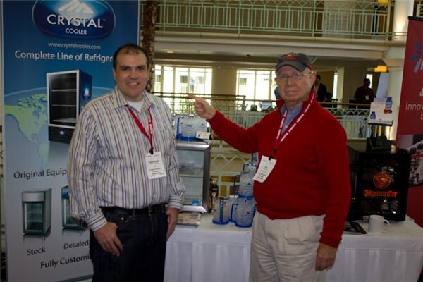 Two men stand at a Crystal Cooler booth; the older man in a red sweater points toward a table with water pitchers and a Jägermeister tap machine.