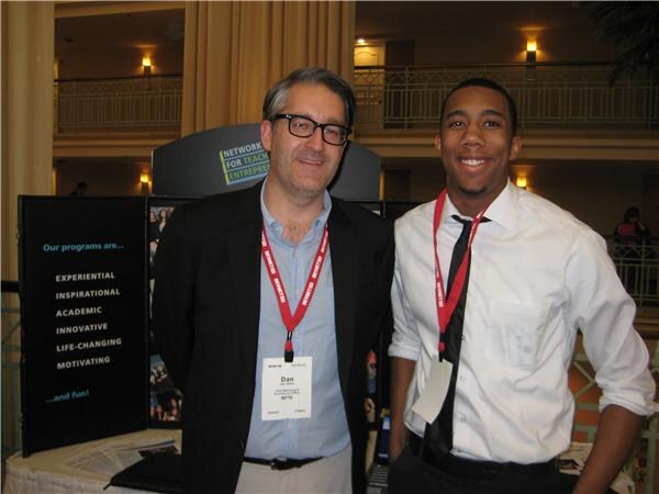 Dan of NFTE, left, in glasses and a dark jacket with a lanyard, stands beside a smiling young man in a white shirt and tie in front of an NFTE display.