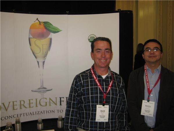 David of Sovereign Flavors, center, wears a plaid shirt and badge in front of a Sovereign Flavors display; a man in glasses stands at right.