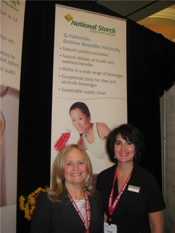 Dinah, right, and a blonde woman smile in front of a National Starch Food Innovation banner for Q-Naturale, both wearing red lanyards.