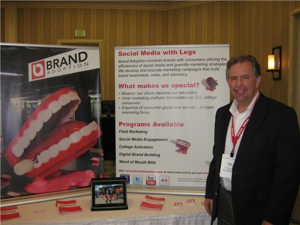 Don, wearing a red lanyard and dark jacket, smiles beside a Brand Adoption booth with banners showing chattering teeth toys and a Social Media with Legs panel.