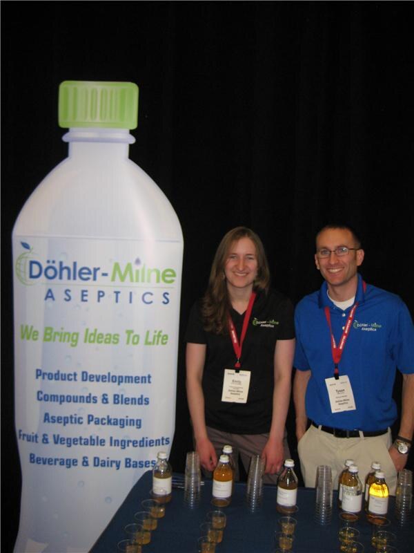 Emily and Tyson stand behind a table of small juice bottles and sample cups beside a large Döhler-Milne Aseptics bottle-shaped banner, both wearing company shirts and red lanyards.