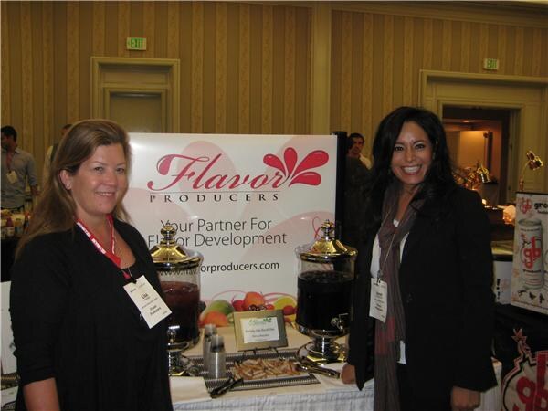 Two smiling women wearing badges stand on either side of a Flavor Producers sign at a trade-show table with glass beverage dispensers, a tray of food and fruit.