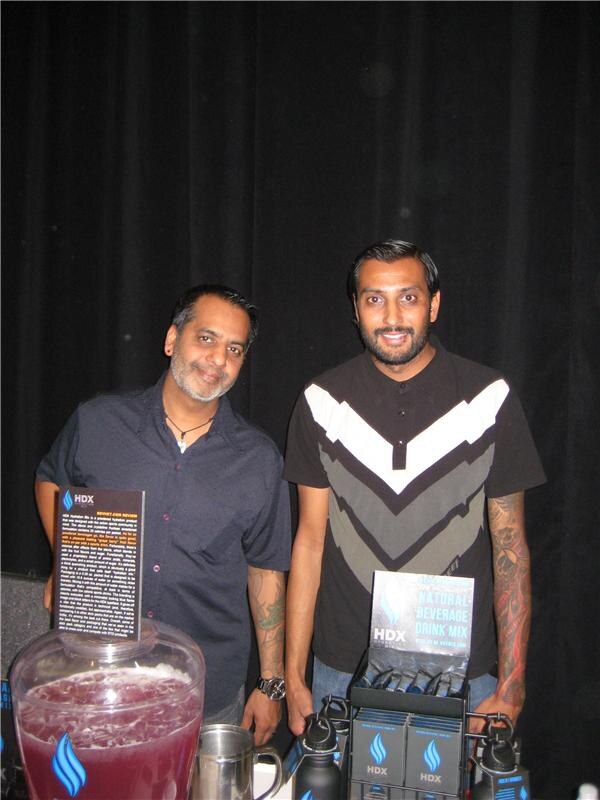 Two men stand behind an HDX display with a jar of purple drink, a printed sign, boxes of drink mix packets and black bottles in front of a dark curtain.