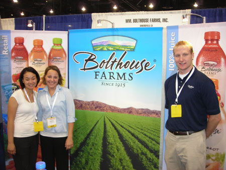 Two women and a man in lanyards stand in front of a Bolthouse Farms banner showing carrot fields, with Bolthouse juice bottles on panels beside them and a Wm. Bolthouse Farms, Inc. sign above.