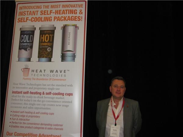 A man wearing a badge stands beside a tall Heat Wave Technologies banner advertising self-heating and self-cooling beverage cups, in front of a dark curtain.